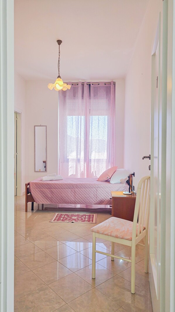 Un dormitorio con cama, una silla, una mesita de noche, un espejo y una ventana con cortinas.