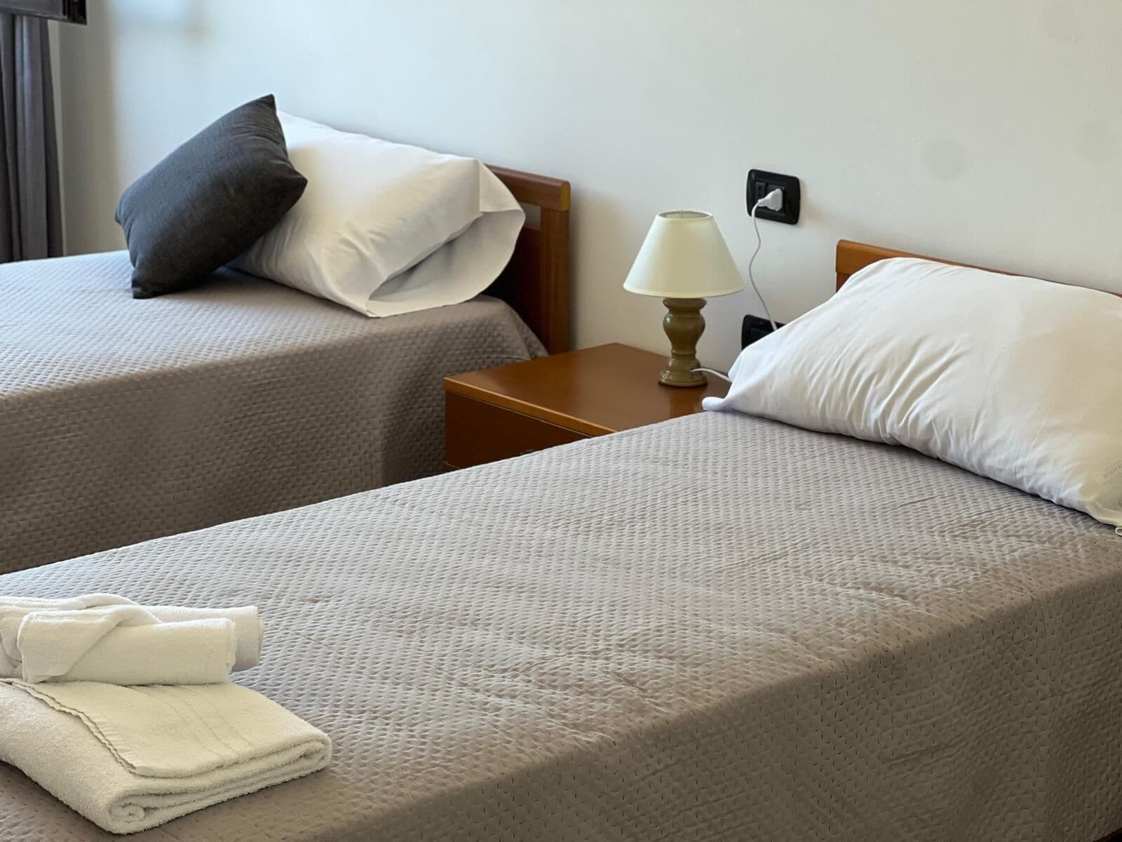 Duas camas de solteiro com roupa de cama cinza texturizada, uma mesinha de cabeceira com abajur e uma tomada na parede.