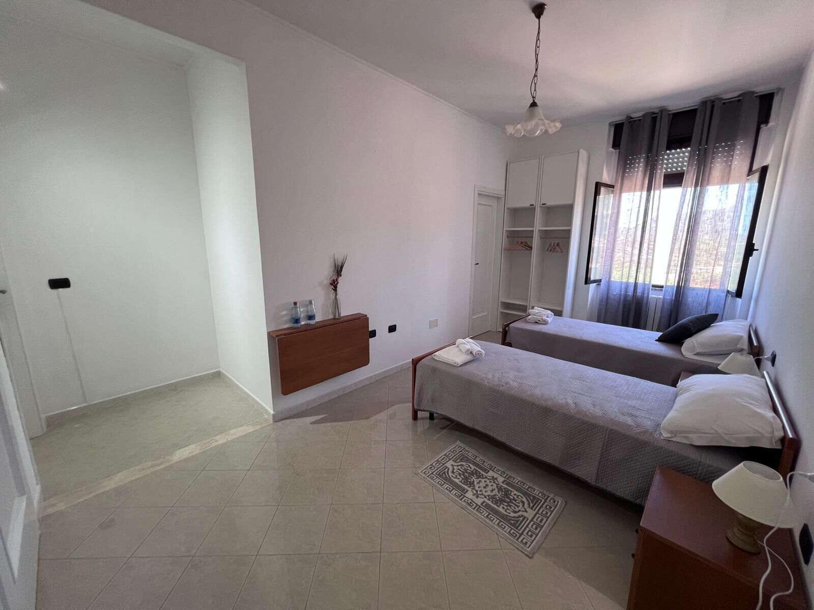 Una habitación de hotel moderna con dos camas individuales, un aparador de madera, una mesita de noche con lámpara, un espejo grande y un armario.