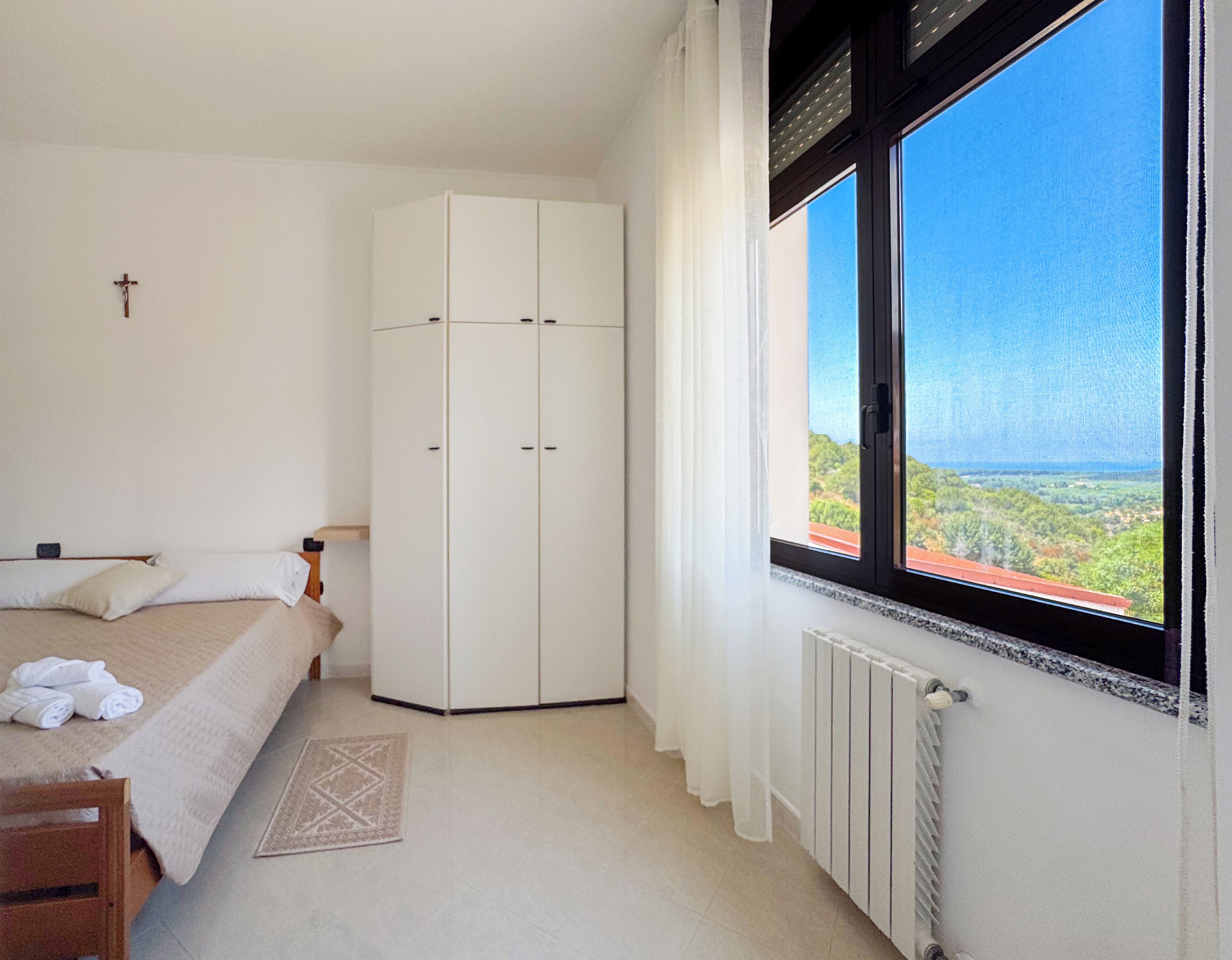 A bedroom with a bed, a wardrobe, a radiator, and a window with a view of greenery.