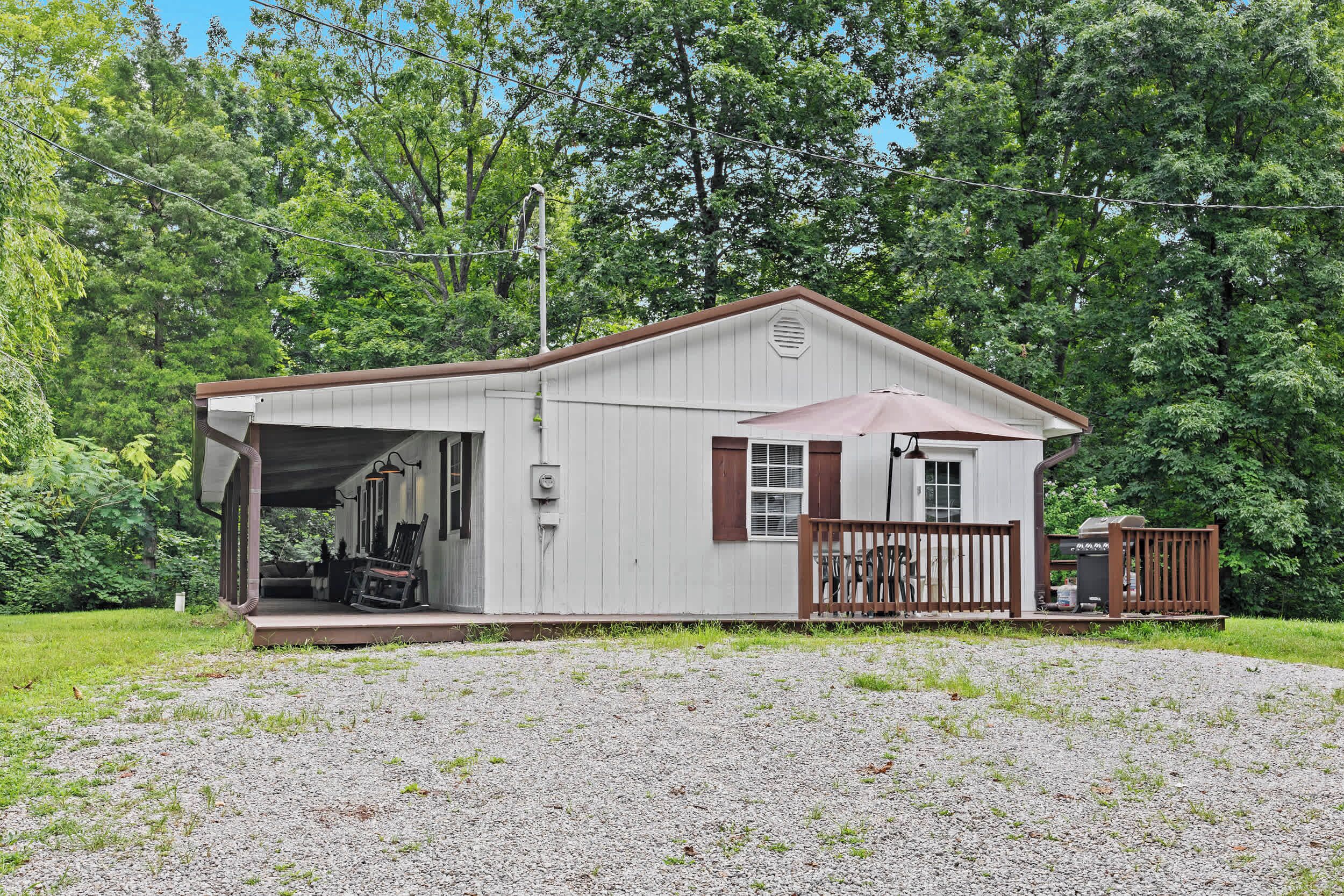A single-story cabin with a covered porch, a small deck, and a gravel driveway.