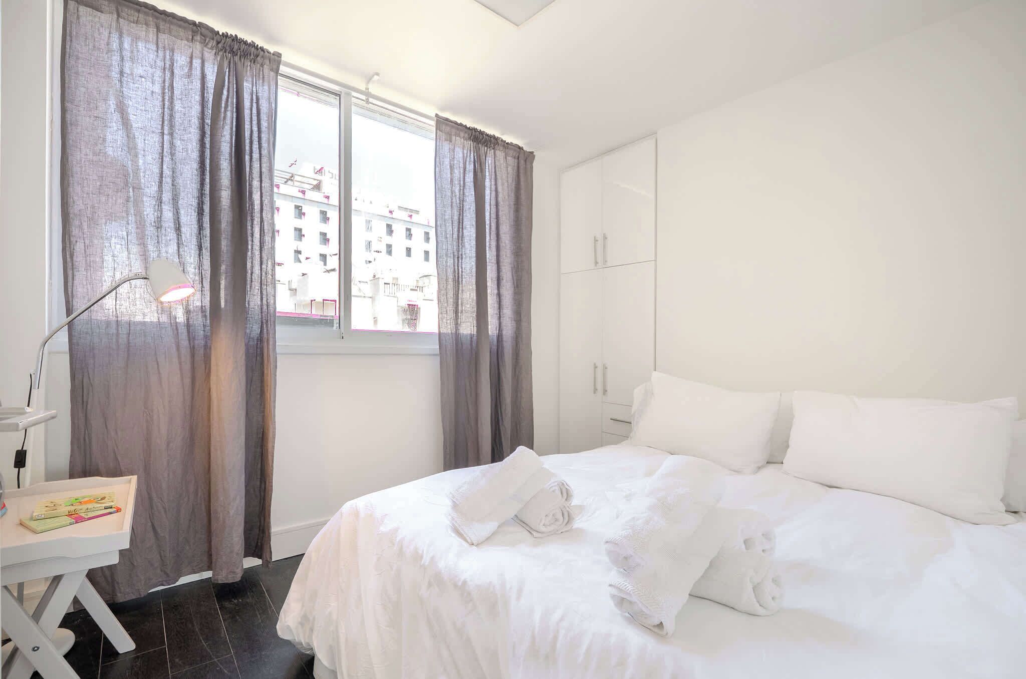 Een schone, witte hotelkamer met een bed, een bureau en een raam met gordijnen.