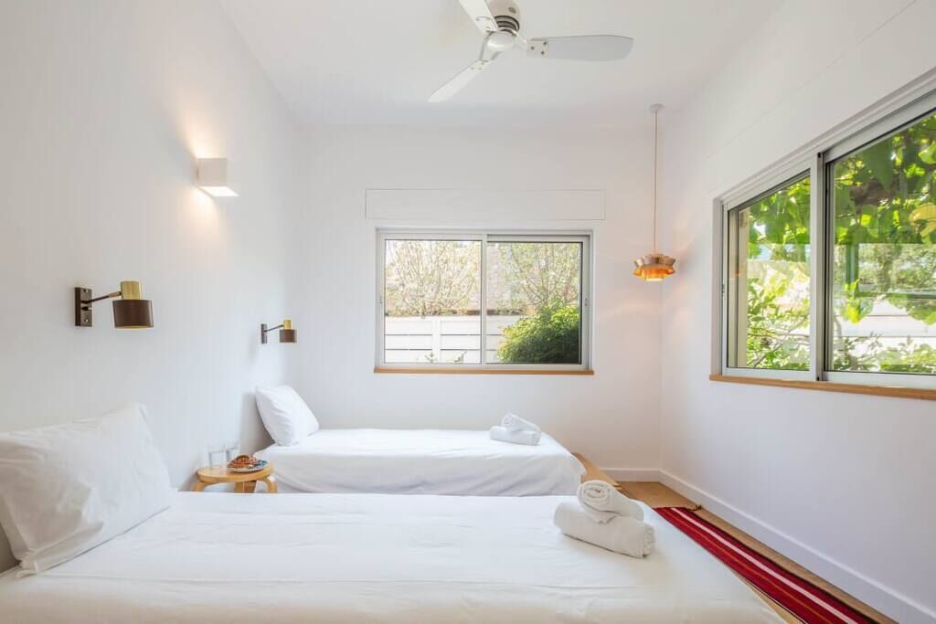 Una camera d'albergo con due letti, una ventola a soffitto, una finestra con vista sul verde e un tavolino con una lampada.