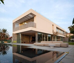 A modern two-story house with a large glass facade, a swimming pool, and outdoor seating.