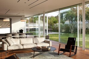 A modern living room with a sofa, a coffee table, and a chair, large glass windows, and a view of a garden.