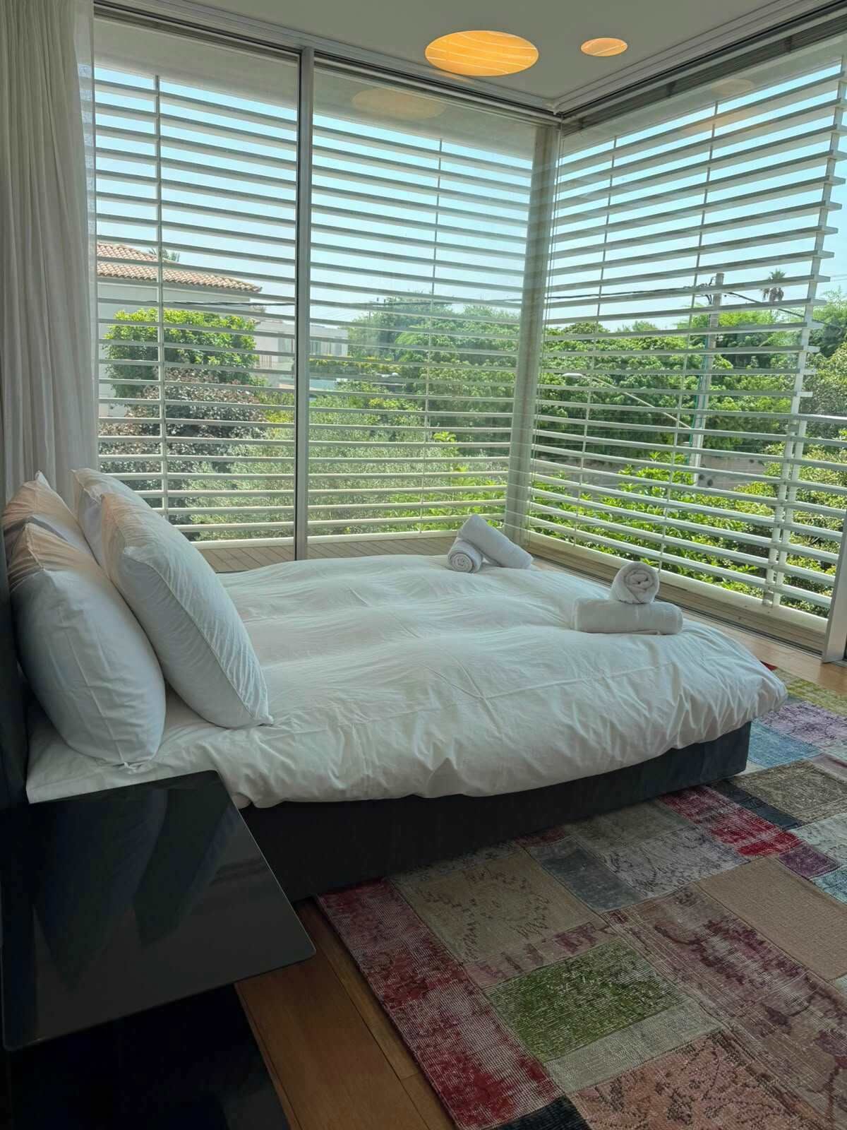 Une chambre moderne avec un grand lit, du linge de lit blanc et une vue sur la verdure grâce à de grandes fenêtres du sol au plafond.