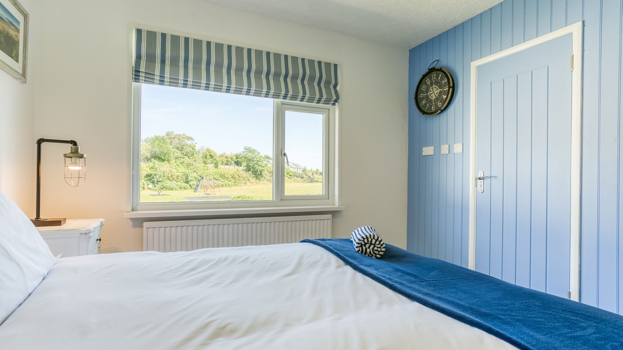 A bedroom with a bed, a window with a view of greenery, a clock on the wall, and a door.