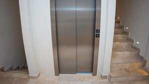 Un ascenseur aux finitions métalliques, un escalier avec des marches de couleur beige et un luminaire fixé au mur.