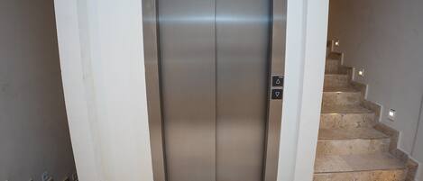 An elevator with a metallic finish, a stairwell with beige steps, and a wall-mounted light fixture.