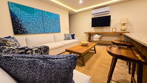 A modern living room with a white sofa, a coffee table, and a large abstract painting. - Cozumel NEW Siesta Studio: 1BR/Wi-Fi/AC/Noise Proof/With View (Cozumel)