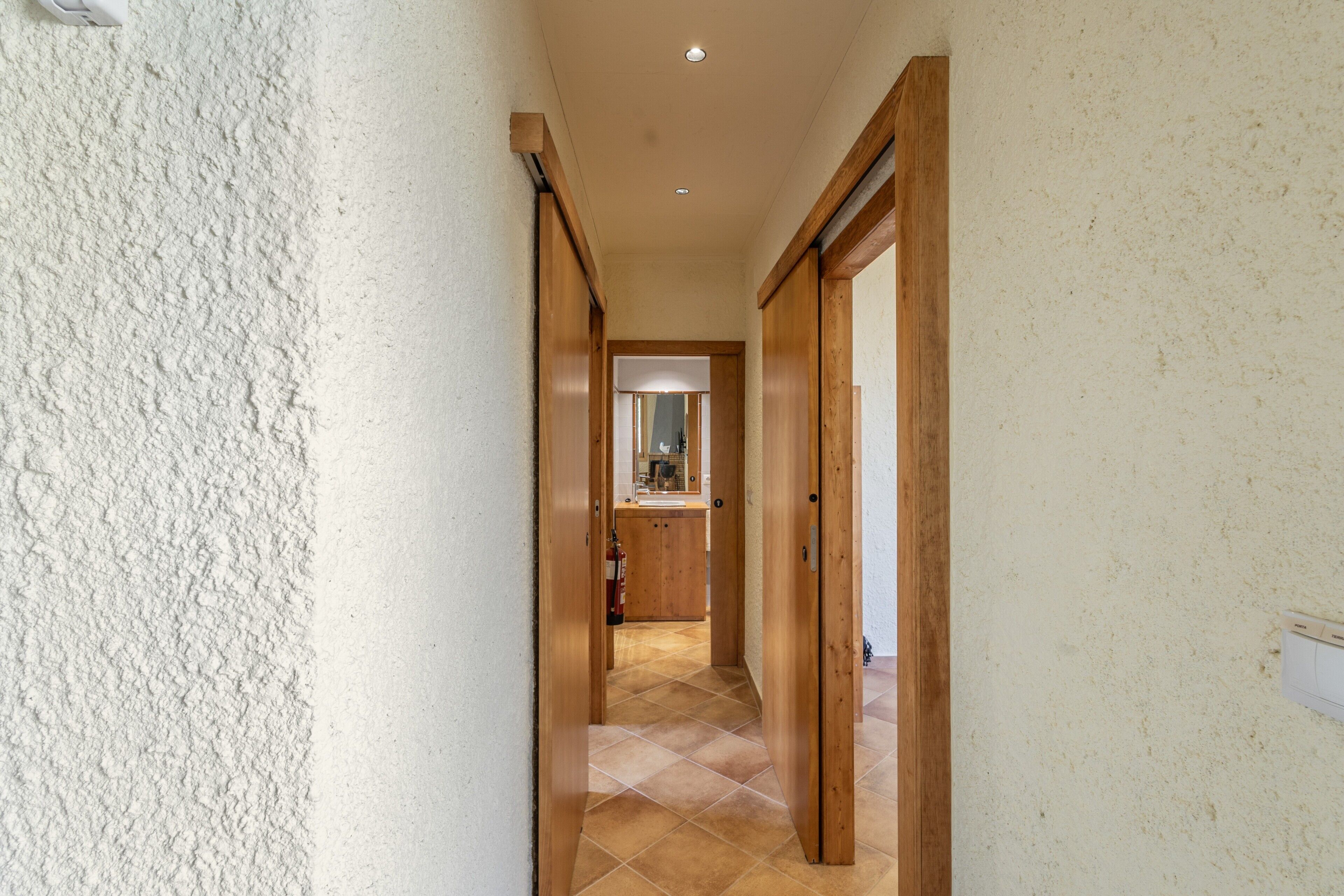 A narrow hallway with a tiled floor, wooden doors, and a visible kitchen area in the distance.