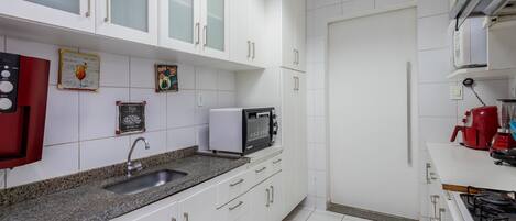Una cocina con gabinetes blancos, encimera de granito, fregadero, microondas y una cafetera roja.