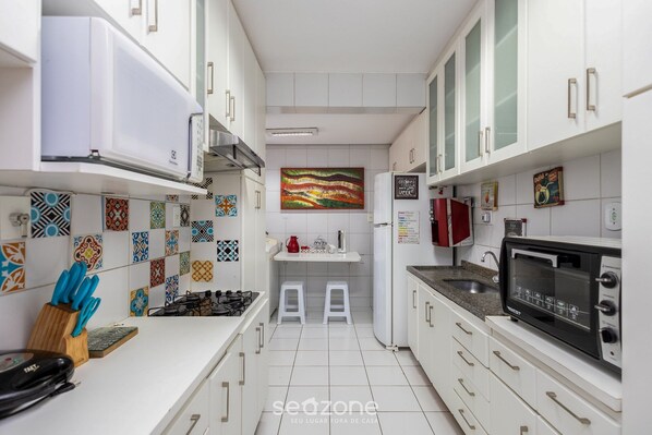 A modern kitchen with white cabinets, a microwave, a gas stove, and a red kettle. There are two stools and a colorful abstract painting on the wall.