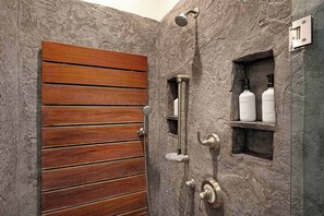 A bathroom with a wooden door, a shower with a handheld showerhead, and two bottles of shampoo and conditioner on a shelf.
