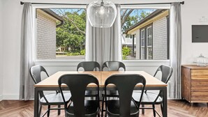 Un coin repas avec une table en bois et des chaises noires, un luminaire suspendu et une vue sur une maison à travers de grandes fenêtres.