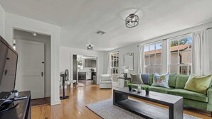 Un salon spacieux avec un plancher en bois, un canapé vert, une table basse et une cuisine à l’arrière-plan.