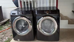 Dos lavadoras de carga frontal ubicadas fuera de una casa con valla de piquete blanca y una puerta.