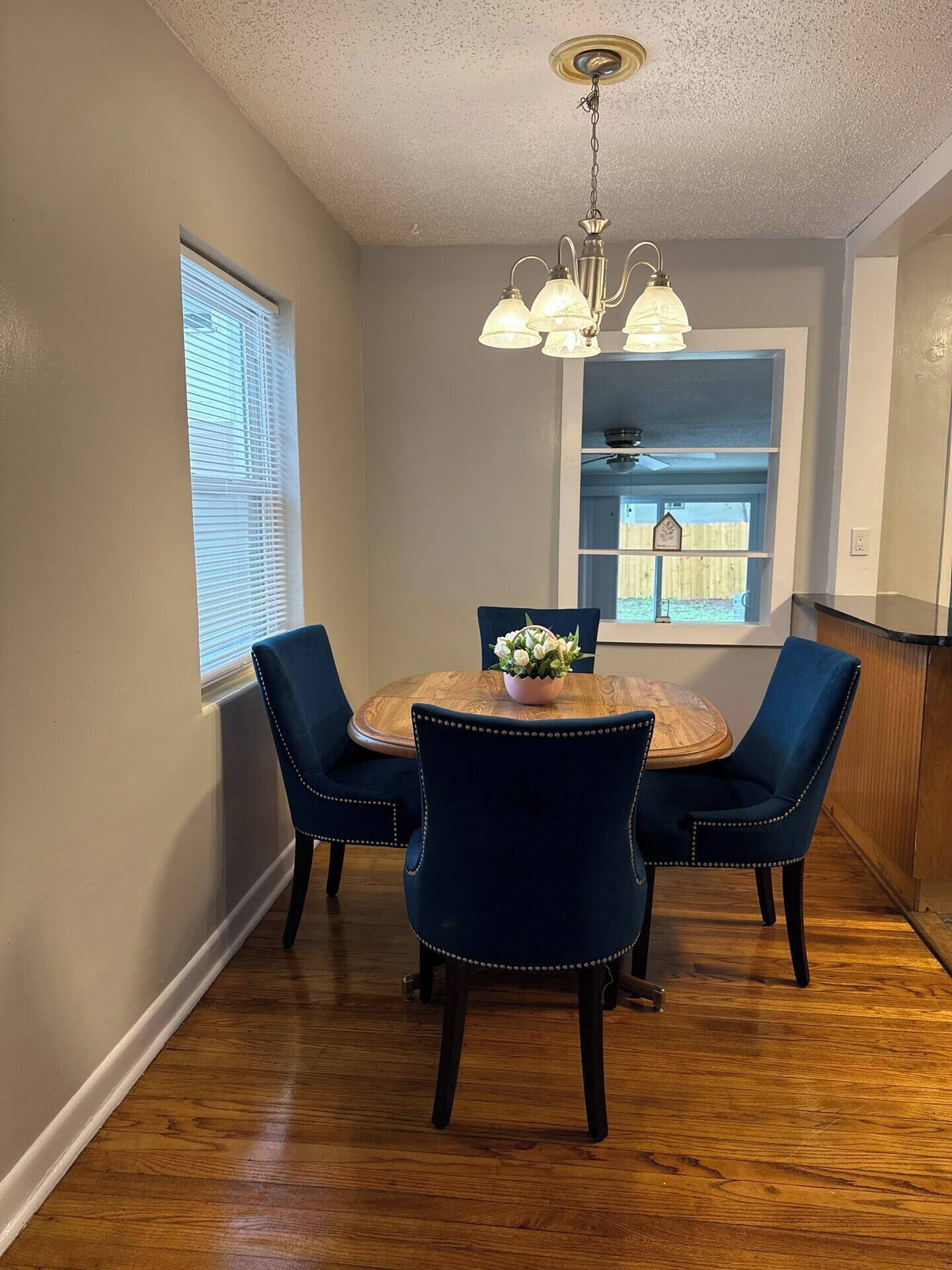 Un coin repas comprenant une table ronde en bois, quatre chaises à l’assise rembourrée de couleur bleu foncé, un lustre et une fenêtre équipée de stores.
