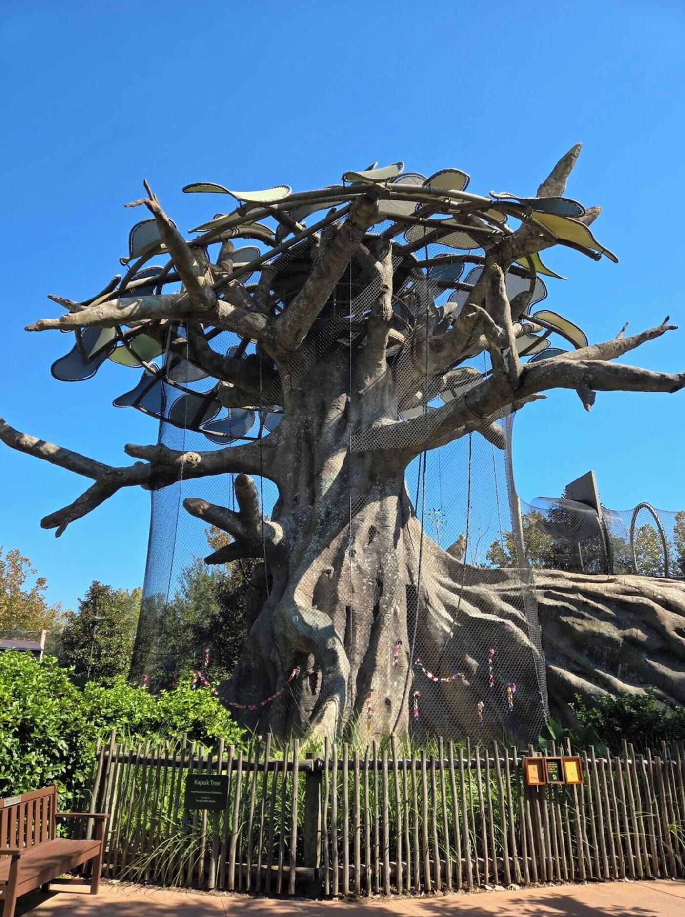 A large tree sculpture with a mesh enclosure, surrounded by a bamboo fence, and a wooden bench in the foreground.