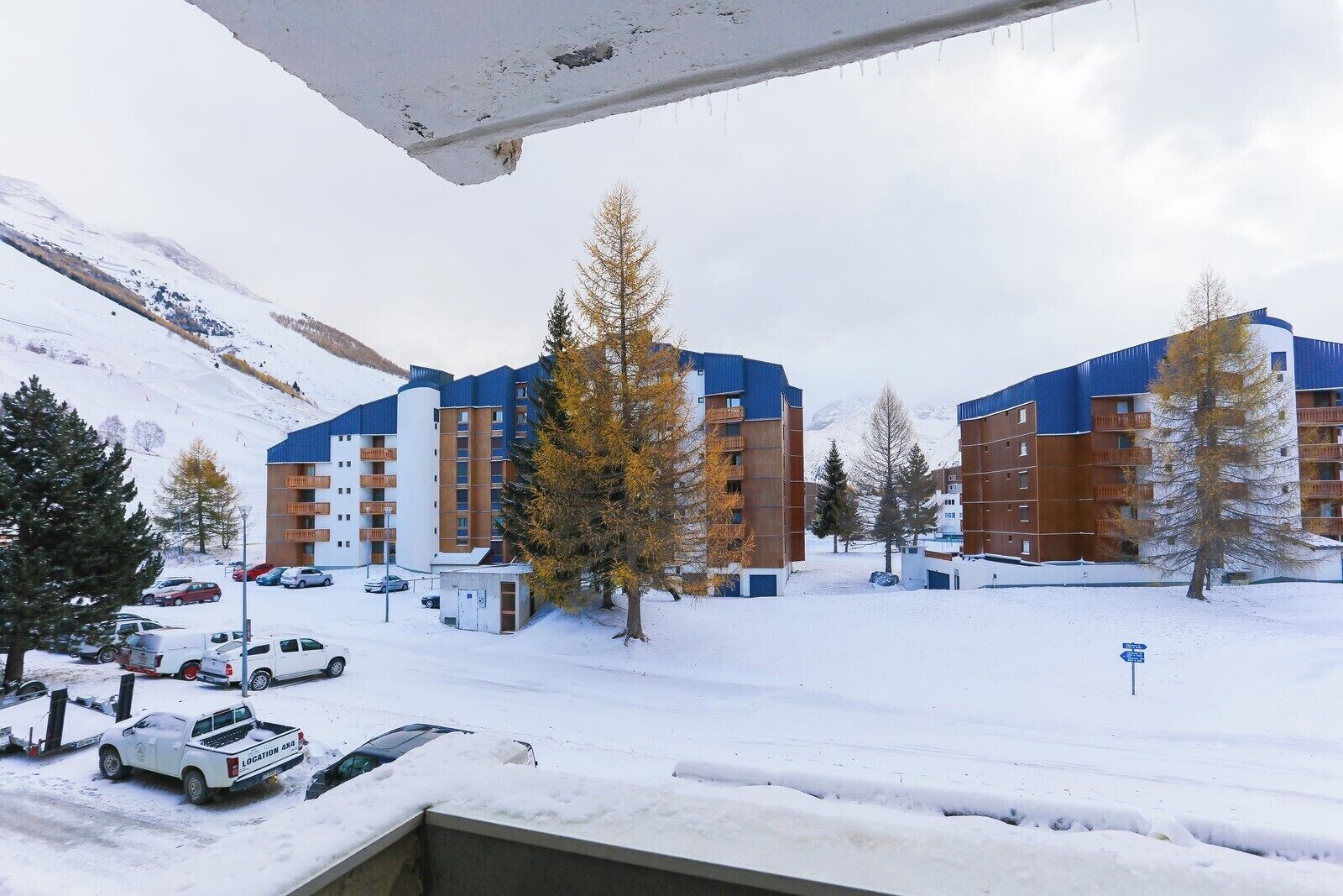 A snow-covered parking area with several cars, a building with balconies, and a mountainous backdrop.