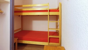 A bunk bed with a ladder, red mattress, and wooden frame in a room with white walls and a door.
