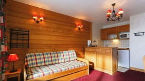Acogedor cuarto tipo cabaña con sofá de madera, una mesita con lámpara, kitchenette con armarios de madera y alfombra roja estampada.
