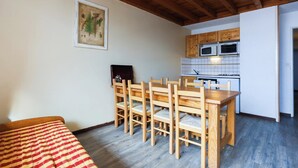 Una habitación compacta con una mesa y sillas de madera, una kitchenette con armarios y electrodomésticos blancos, e ilustración botánica enmarcada en la pared.