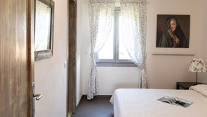 A bedroom with a bed, a window with floral curtains, a nightstand with a lamp, and a painting on the wall. - Terrazza al Poggio Vista Mare by Wonderful Italy (Olbia)