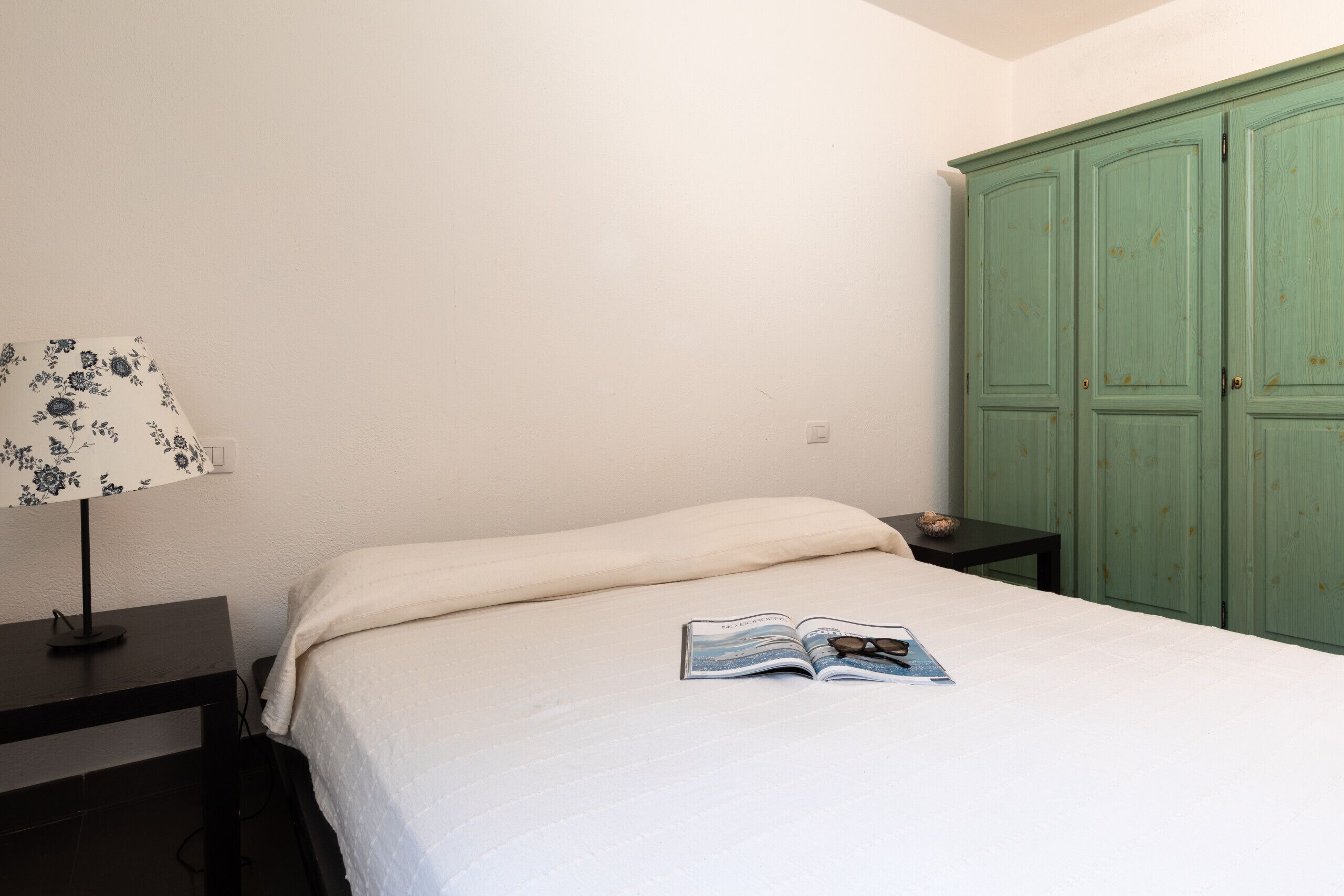 A bedroom with a bed, a nightstand, a lamp, and a green wardrobe.