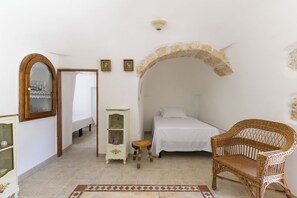 A bedroom with a bed, a wicker chair, a small stool, a cabinet, and an arched doorway. - Trulli Isabella by Wonderful Italy (Ceglie Messapica)