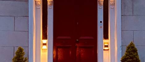 A building entrance with a dark red door, numbered 105, flanked by two potted plants and two wall-mounted lights.