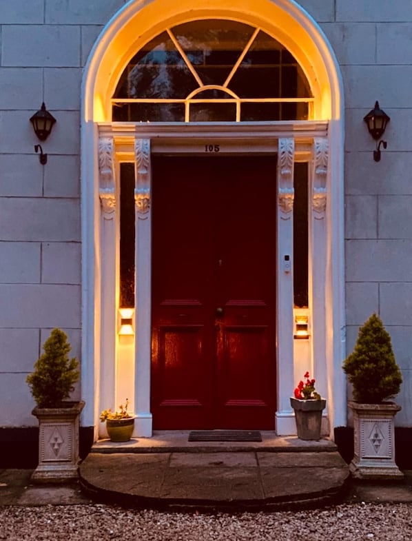 A building entrance with a dark red door, numbered 105, flanked by two potted plants and two wall-mounted lights. - Fabulous 6 Bedroom Victorian Period House (Londonderry)