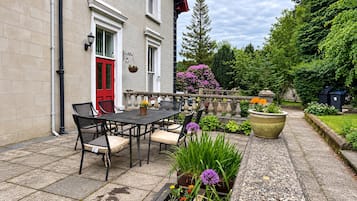 Un patio con mesa y sillas, una puerta roja y un jardín con diversas plantas y flores.
