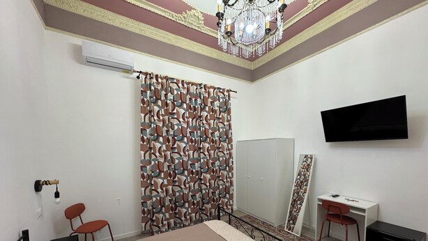 A hotel room with a bed, a desk, a chair, a wardrobe, a TV, and a patterned curtain. - Caltur B&B (Palermo)
