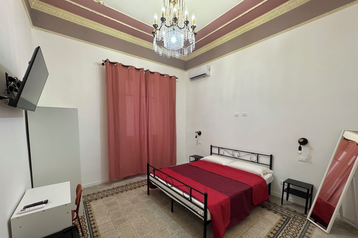 A bedroom with a bed, a desk, a chair, a chandelier, and a mirror.
