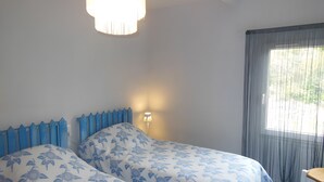 A room with two beds, a window with curtains, a chair, and a wall-mounted light.