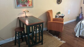 Ein Zimmer mit einem Tisch, zwei Stühlen, einer Lampe, einem Teppich, einer Gitarre sowie einer Wand mit einem Gemälde und dekorativen Tellern.