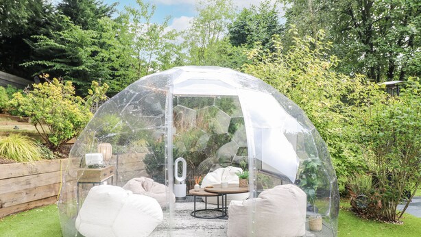 Eine transparente Kuppelkonstruktion mit Sitzgelegenheit innen, inklusive Tisch und Stühlen, umgeben von einem Garten.