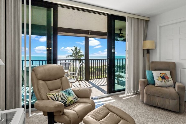 A room with a large sliding glass door leading to a balcony with a view of the ocean and palm trees. There are two leather chairs, one with a pillow and another with a cushion.