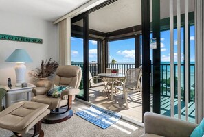 Een balkon met uitzicht op de oceaan, een ronde tafel met stoelen, een beige fauteuil, een tafeltje met een lamp, en een kaart op de vloer.