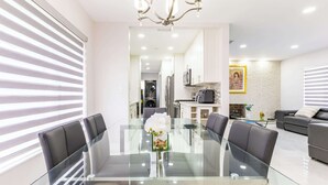 Cozinha moderna com eletrodomésticos de aço inoxidável, mesa de jantar de vidro e sala de estar com sofá.