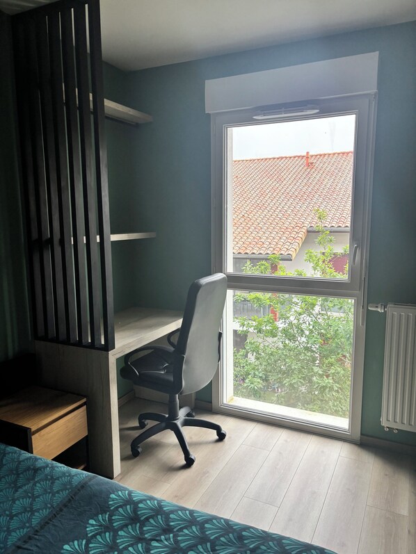 A room with a desk, chair, and a window overlooking a tiled roof and greenery. - Bright apartment 15 minutes from Bayonne (Lahonce)
