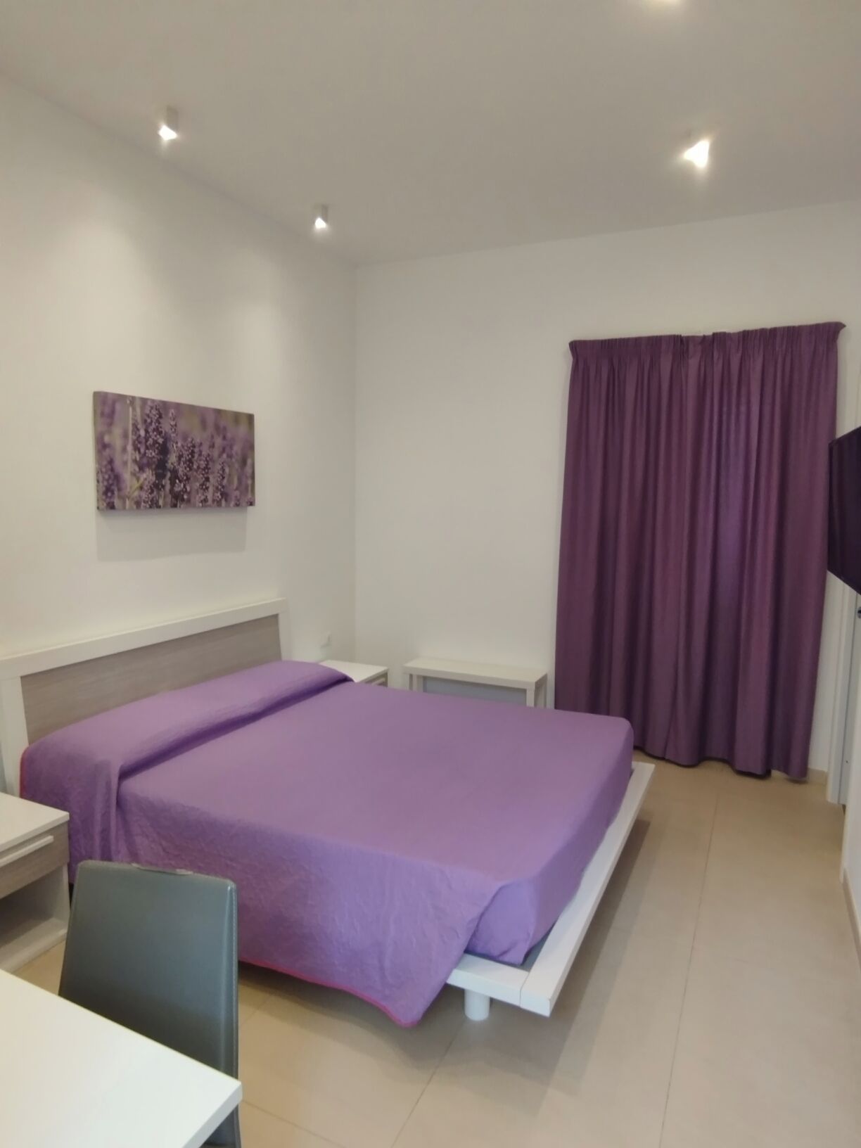 Een slaapkamer met een bed, een bureau, een stoel, een televisie en een raam met gordijnen.