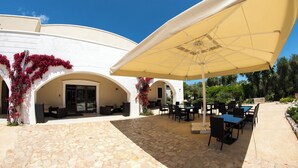 Une terrasse extérieure couverte, dotée d’un sol en pierre, de tables, de chaises et d’un grand parasol.