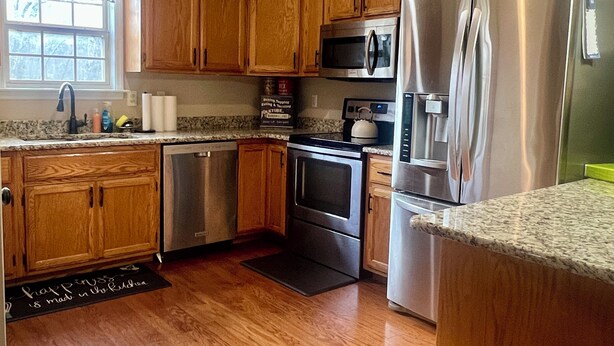 A kitchen with wooden cabinets, stainless steel appliances, a microwave, a refrigerator, a dishwasher, and a window with a view of trees.