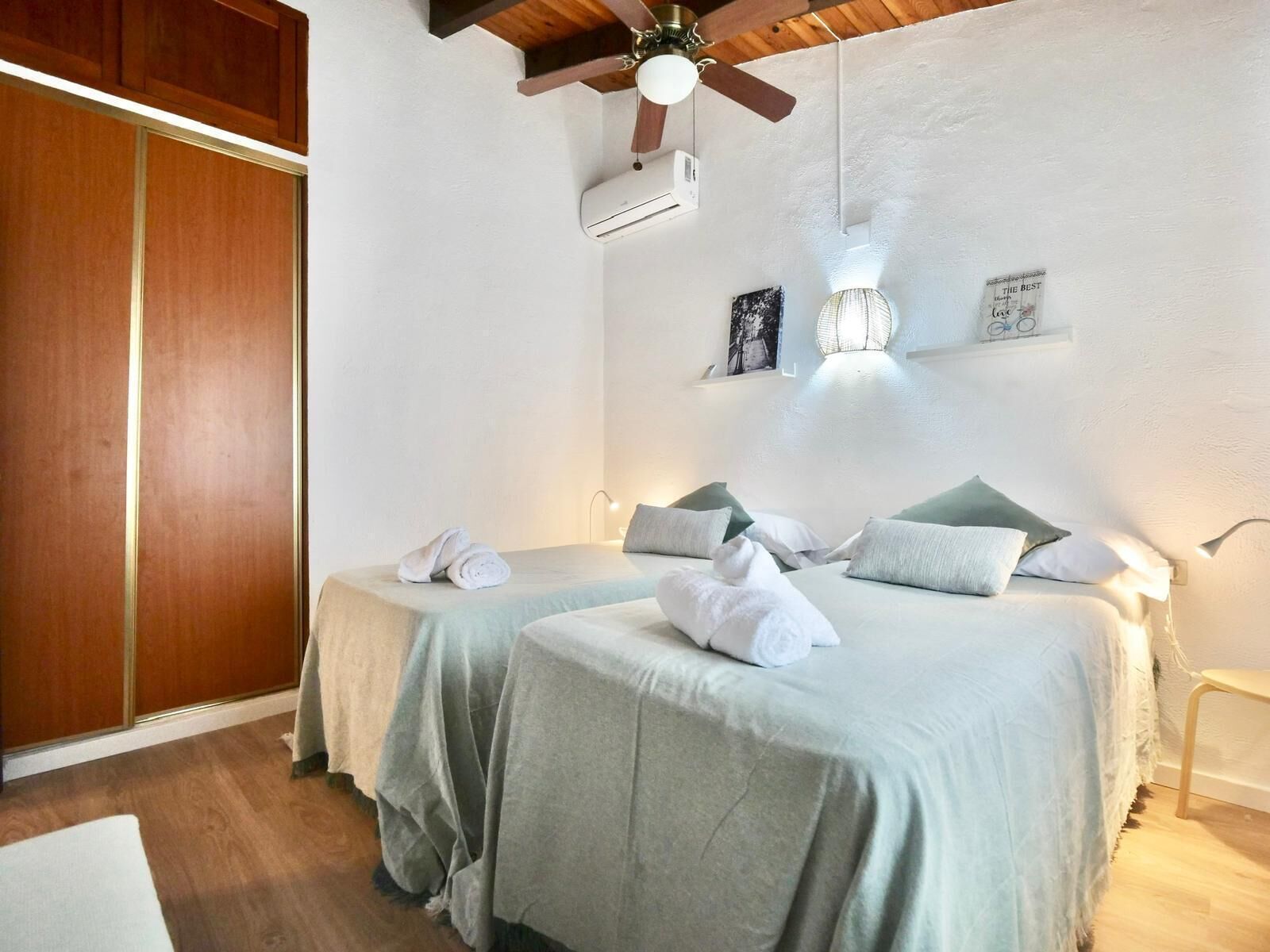 Ein Hotelzimmer mit zwei Betten, einem Holzschrank, einem Deckenventilator, einer Wandklimaanlage und einem kleinen Regal mit Gegenständen.