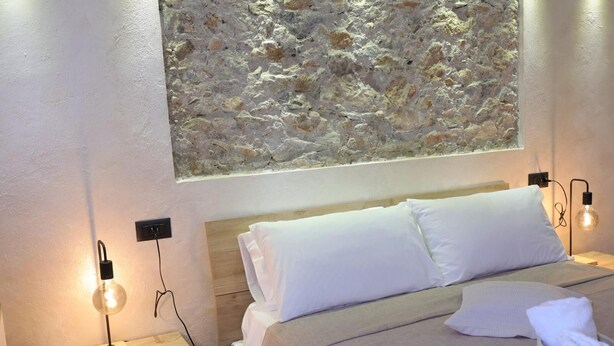Une chambre avec un mur en pierre, une tête de lit en bois, deux tables de chevet avec des lampes et un lit bien fait, recouvert de linge de lit blanc.