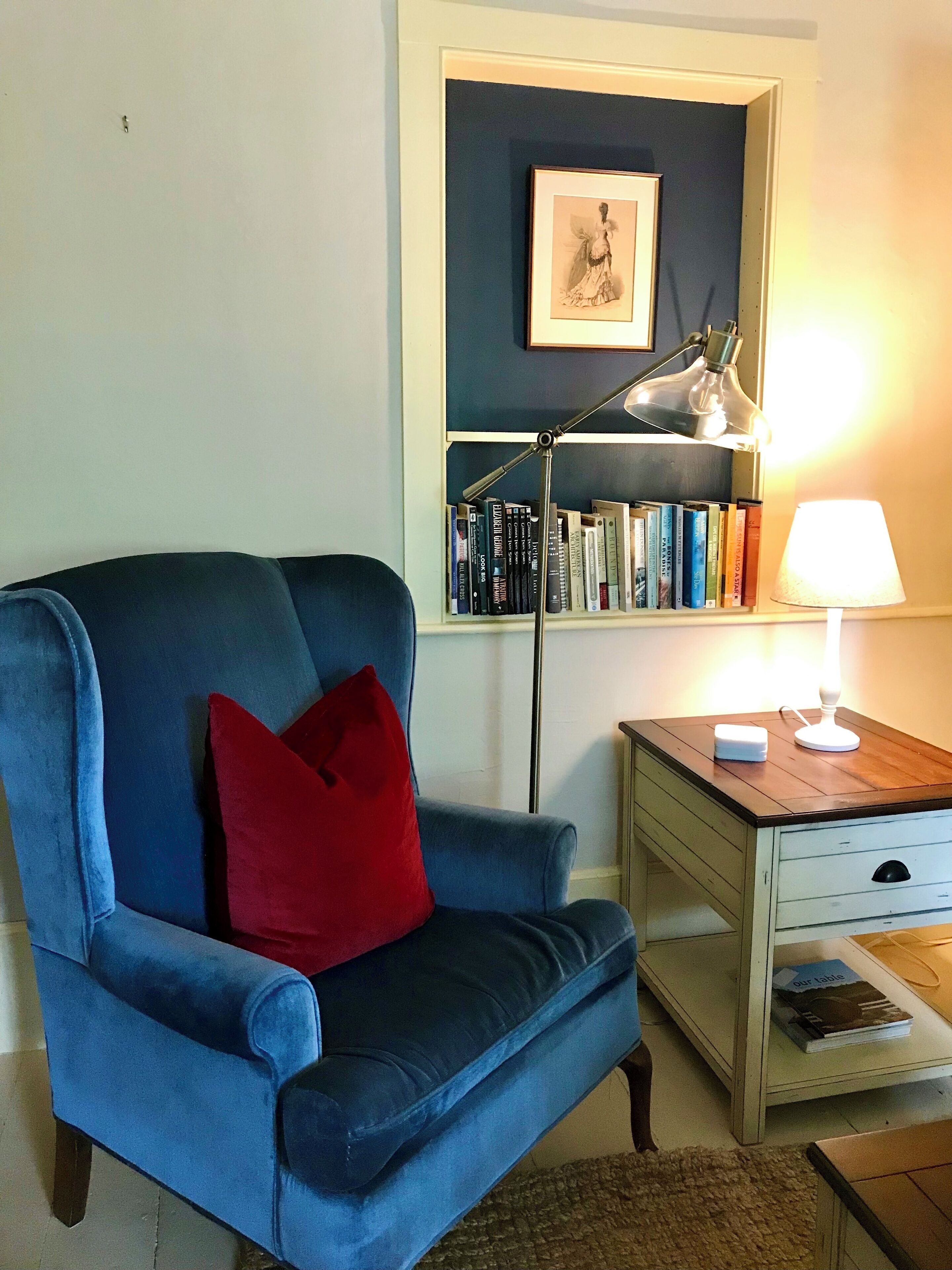 Ein Zimmer mit blauem Sessel, rotem Kissen, Bücherregal, Schreibtisch mit Lampe und einem gerahmten Bild an der Wand.