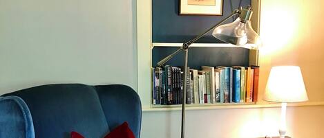 Ein Zimmer mit blauem Sessel, rotem Kissen, Bücherregal, Schreibtisch mit Lampe und einem gerahmten Bild an der Wand.