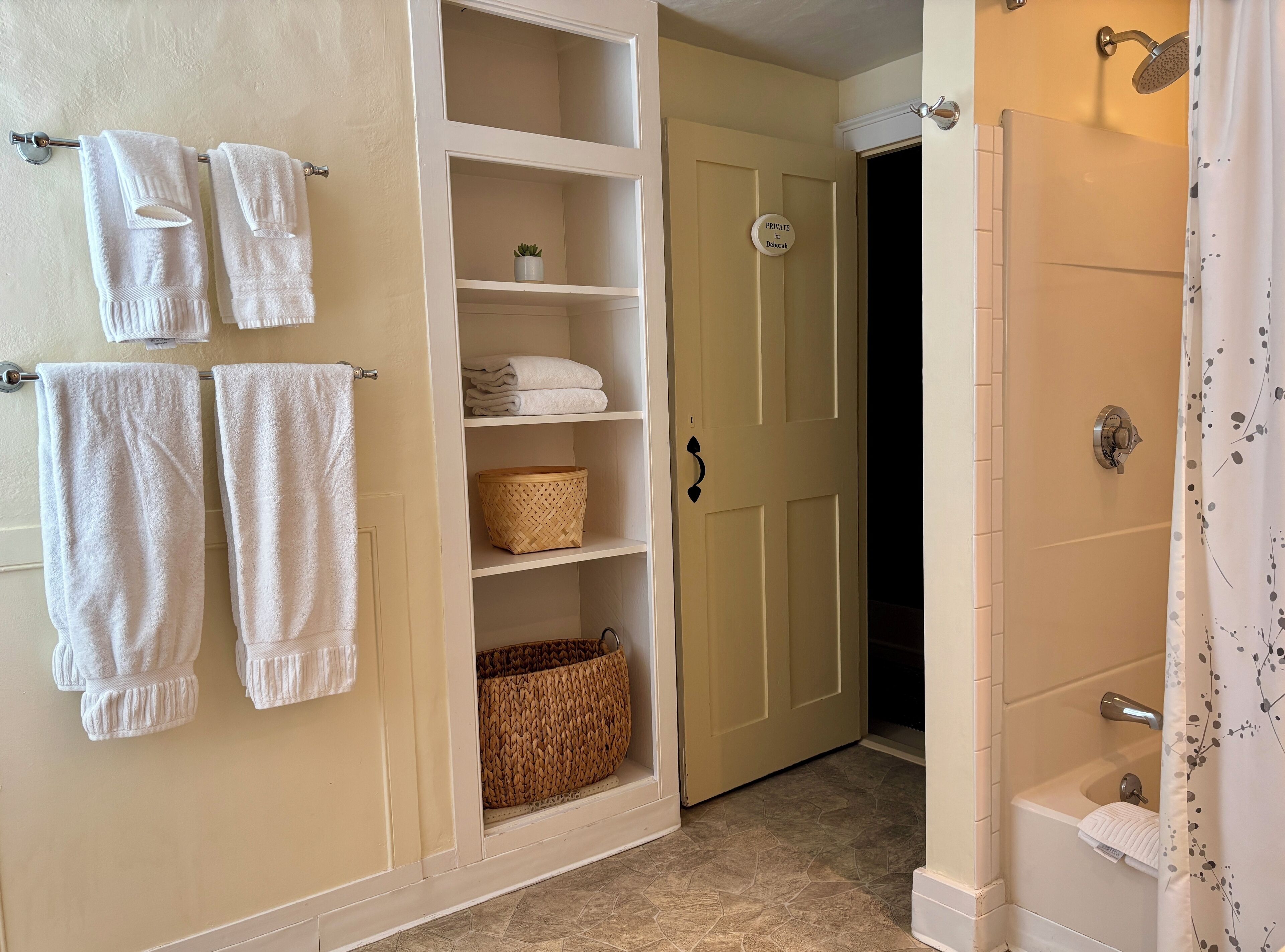 A bathroom with a shower, a towel rack with towels, a storage unit with baskets, and a door labeled "PRIVATE for Deborah".